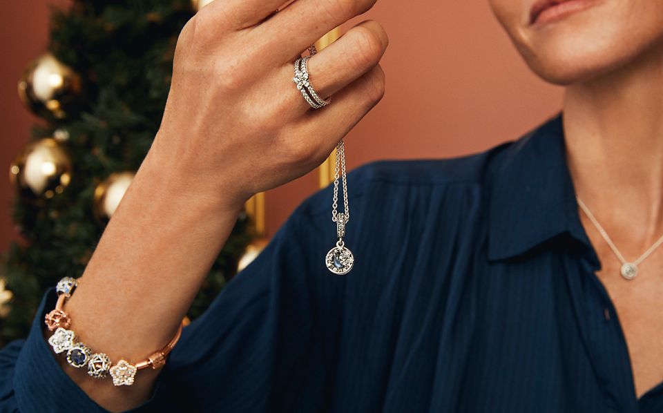 模特佩戴饰有精美串饰的手链及项链，手持 Pandora 项链和戒指。