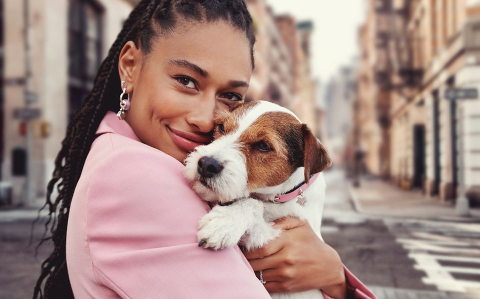 Donna che tiene in braccio un cane che indossa un collare per animali Pandora Moments. Donna che indossa orecchini a cerchio con charm rosa.