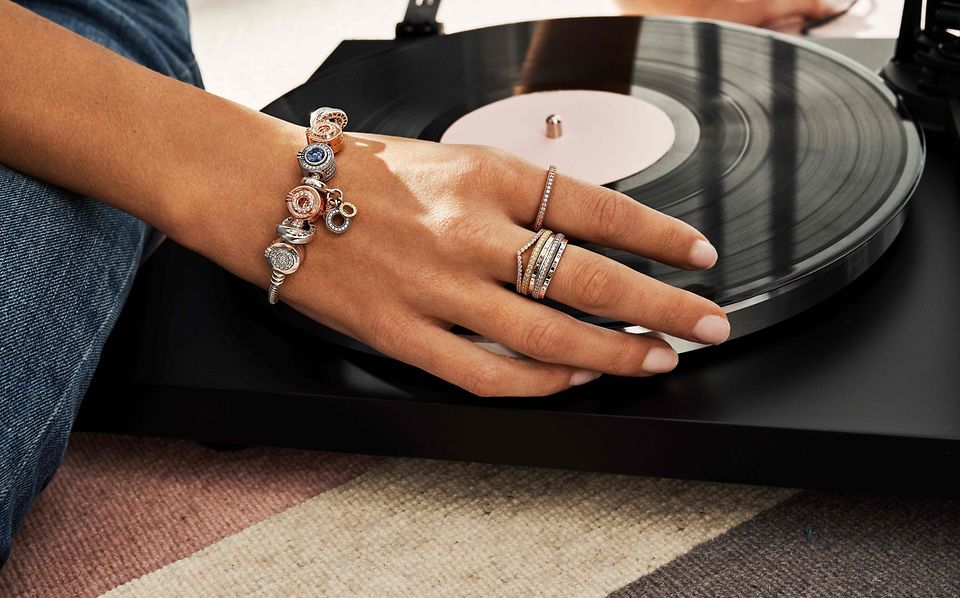 Hånd på vinylplade, der bærer stabelbare ringe og charmarmbånd fra Pandora Signature